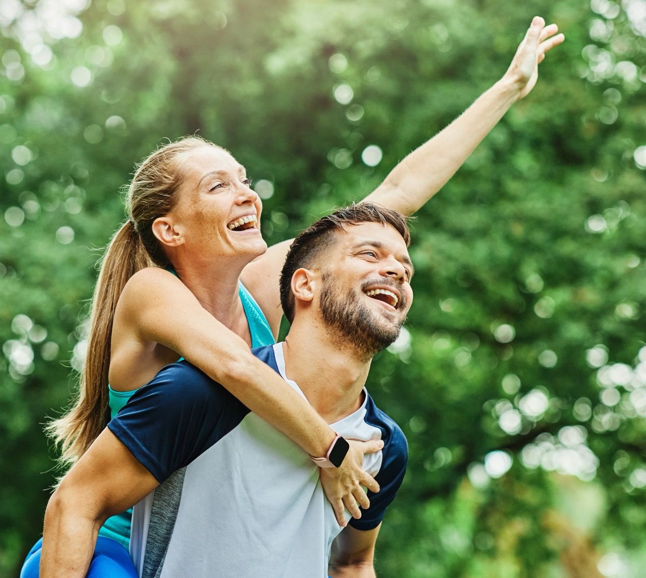 Smiling couple outdoors, the woman riding on the man's back with an arm raised in a green park