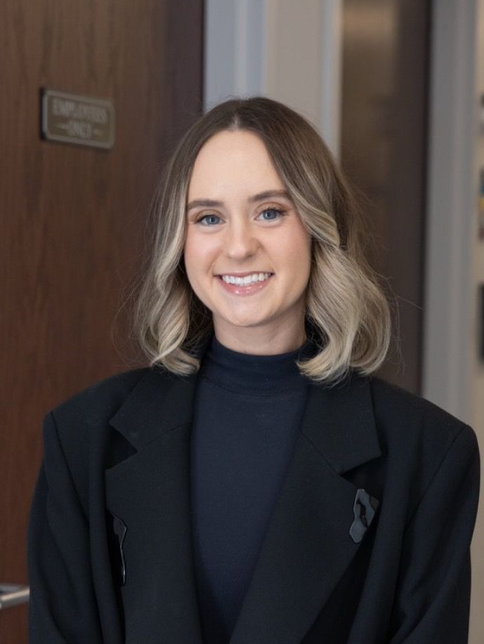 Smiling person in a black blazer and dark top standing indoors by a wooden door