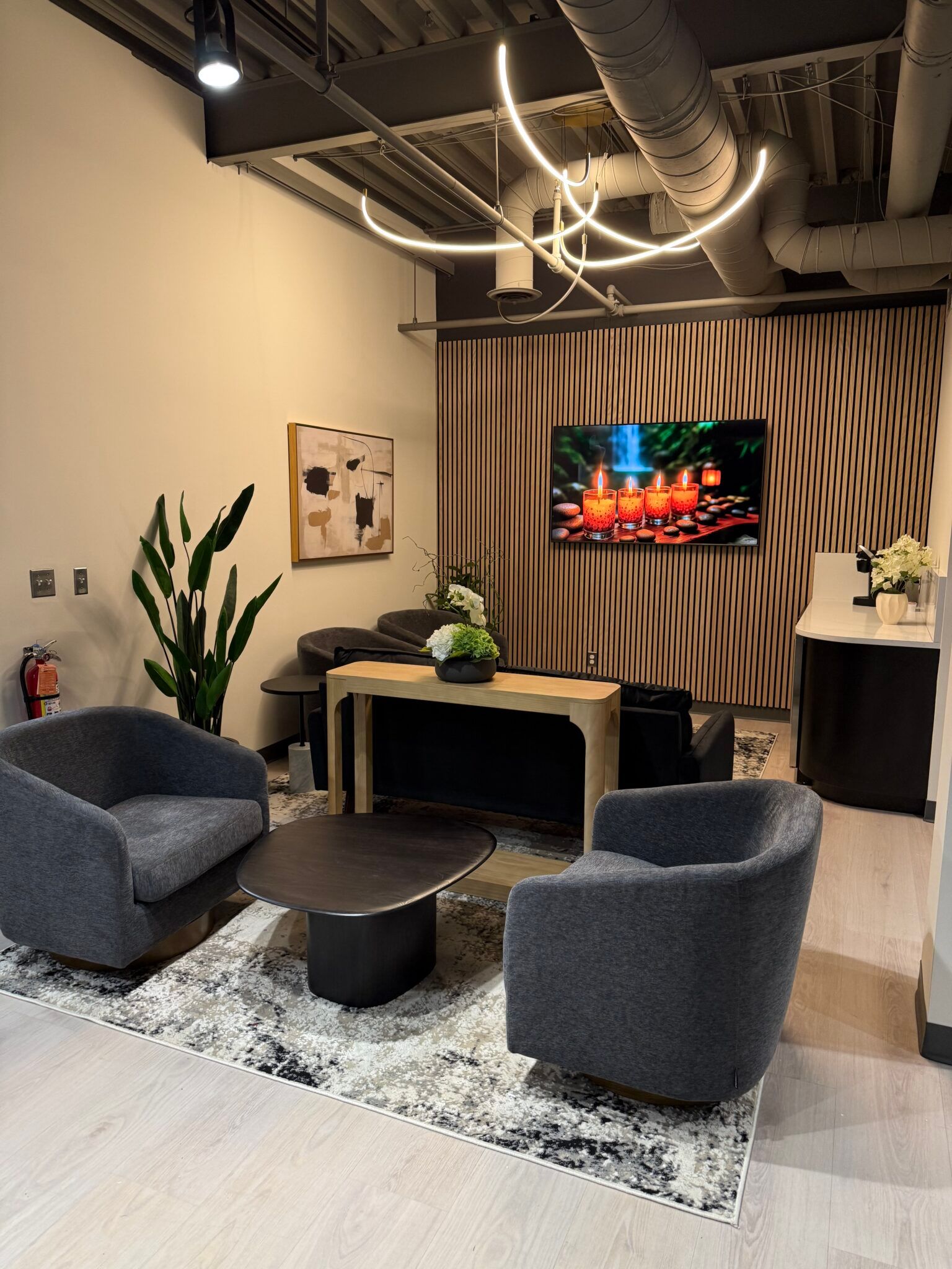 Modern lounge with gray chairs, black tables, TV, plants, and exposed ceiling pipes.