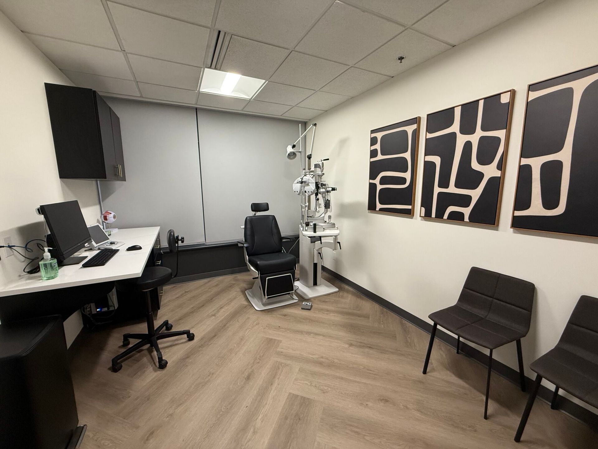Medical exam room with exam chair, wall art, desk, and black waiting chairs on wood floor.