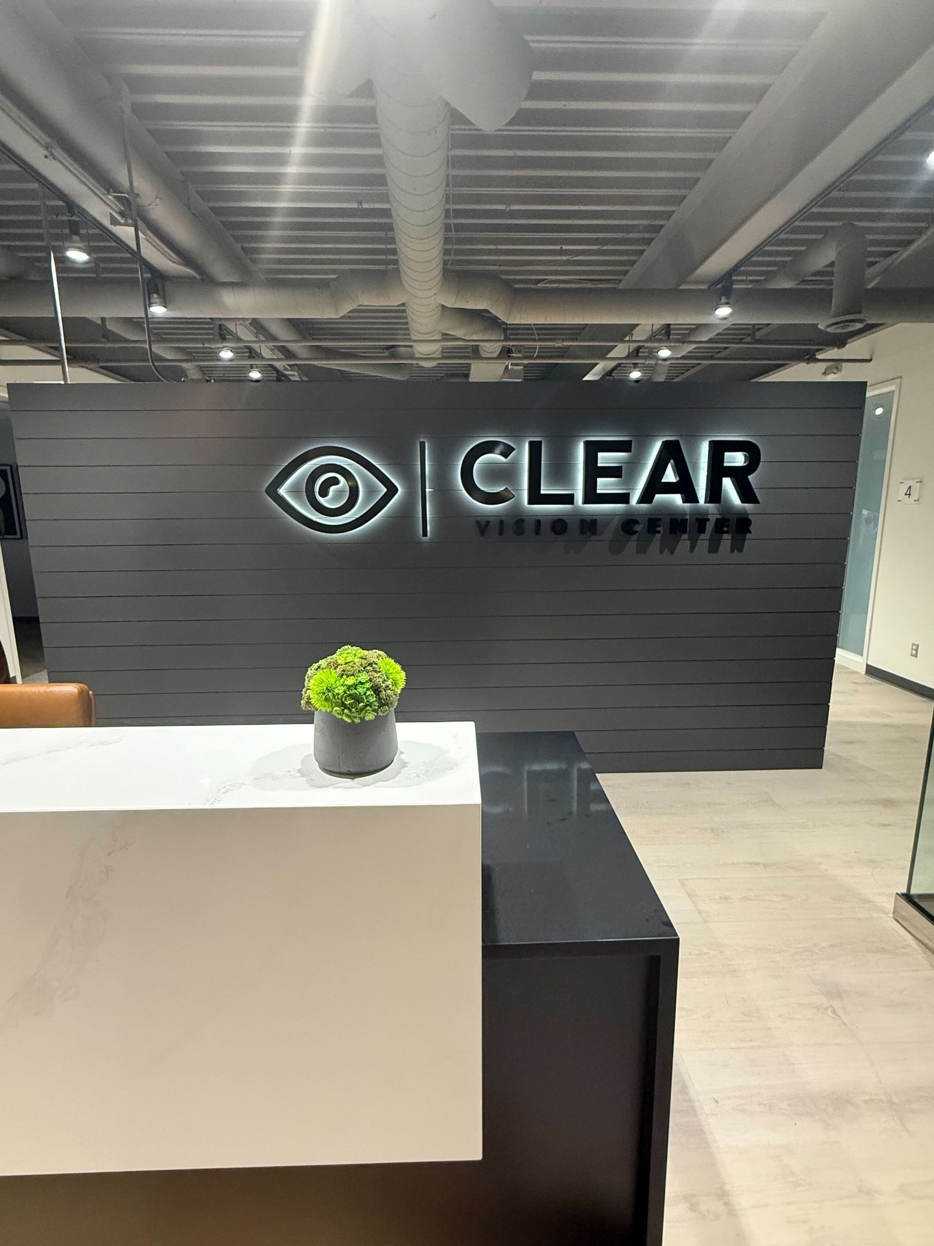 CLEAR office reception area with black wall sign, white counter, and small potted plant
