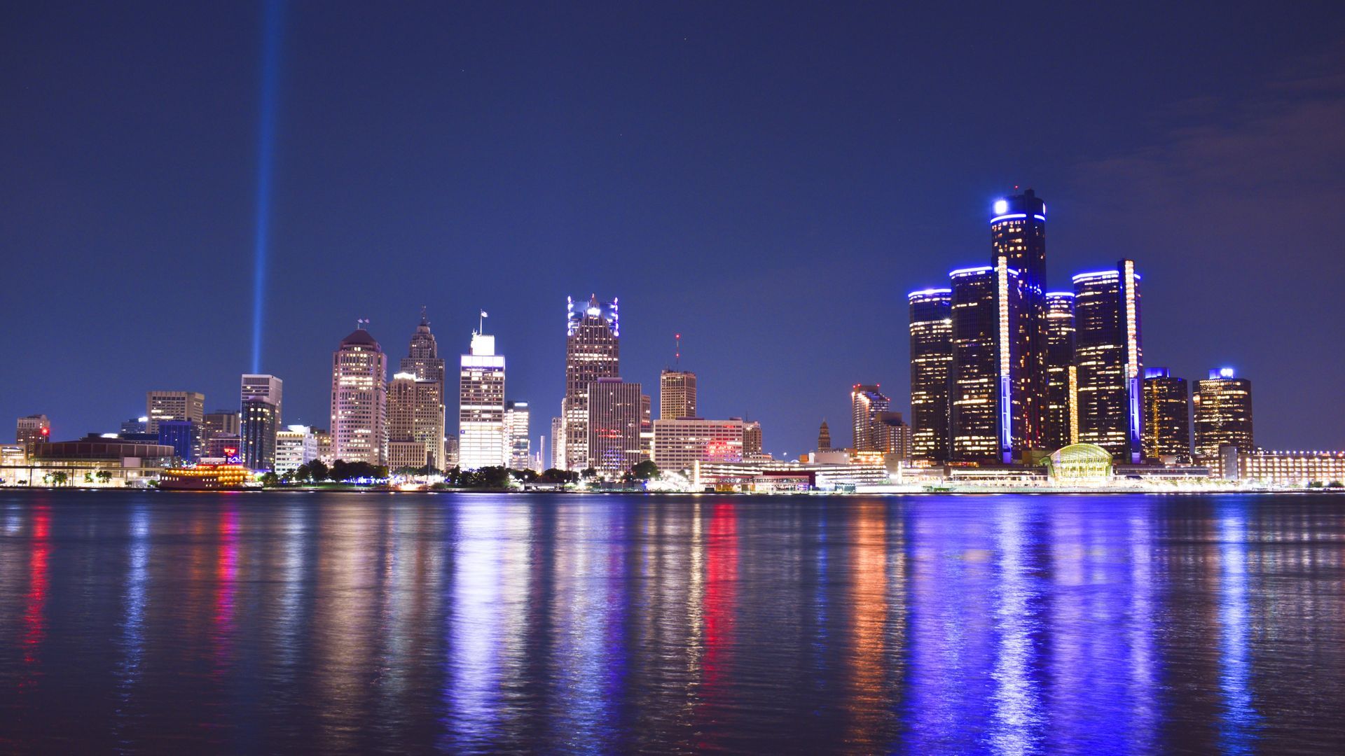 Nighttime skyline of Detroit, Michigan, with city lights reflected in the water. A tall illuminated beam pierces the dark sky.
