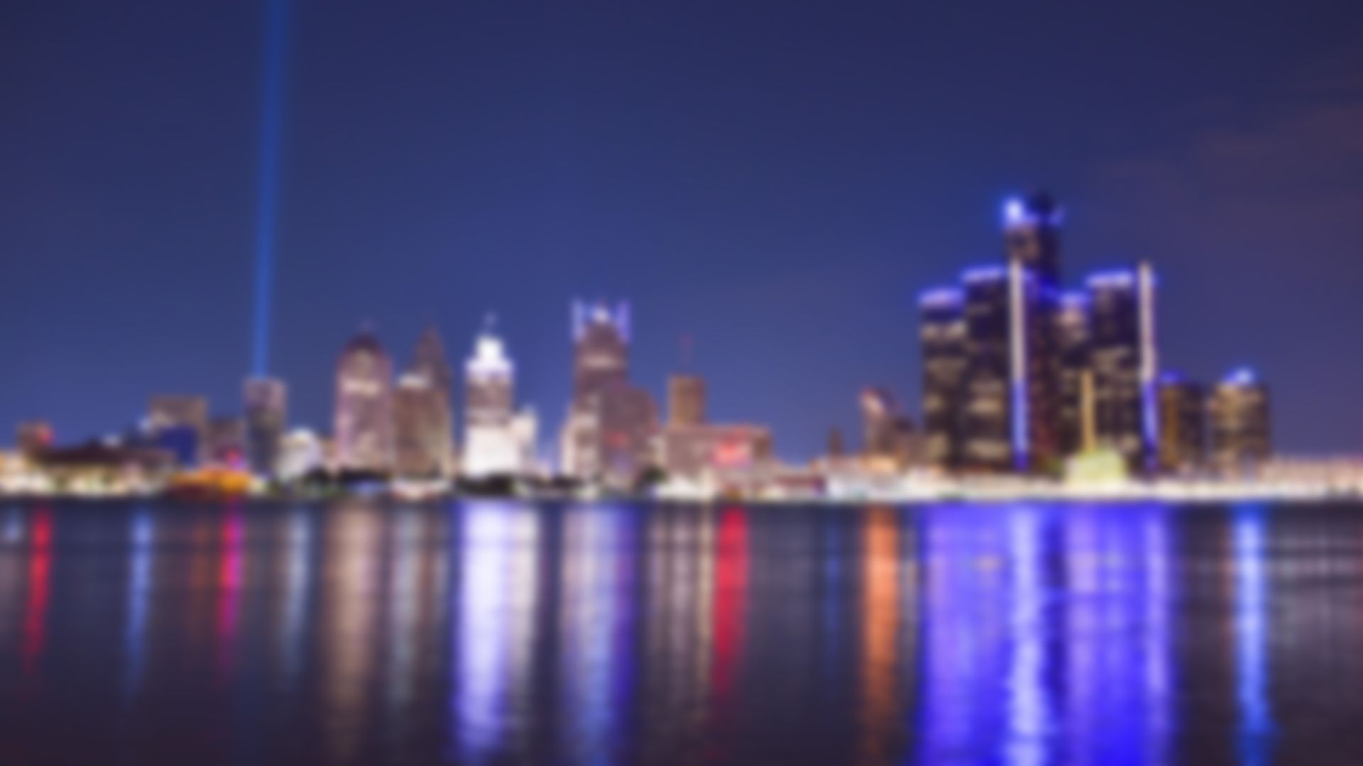 Detroit skyline at night with city lights reflected in the water. A tall beam of light points upward.