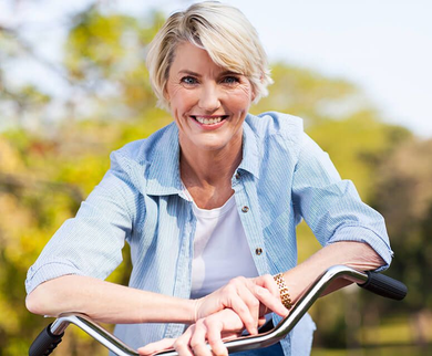 Woman smiling, leaning on bicycle handlebars outdoors; wears blue shirt, white t-shirt, blond hair.