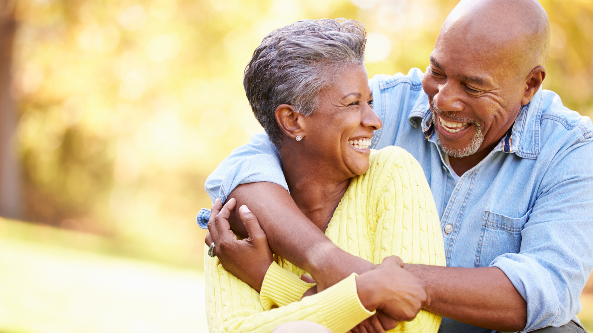 Two smiling people embracing outdoors in warm sunlight, wearing yellow and blue shirts.