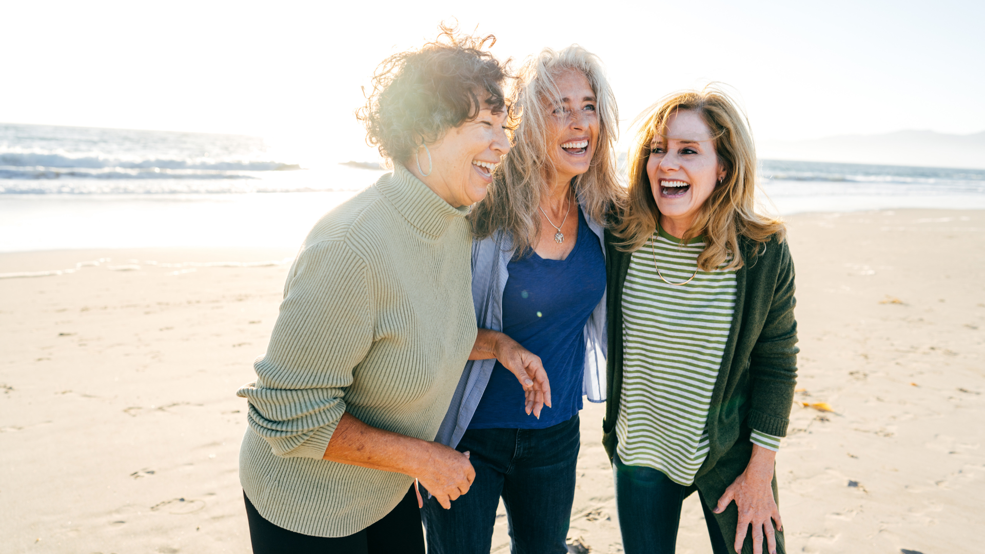 Three people laughing on a sunny beach, with ocean waves in the background.