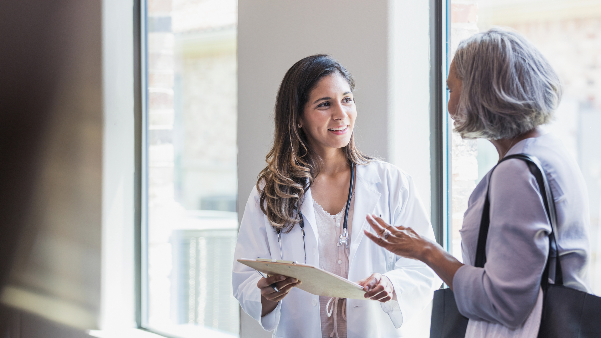 Two women talking in a bright clinic hallway, one in a white coat holding papers.