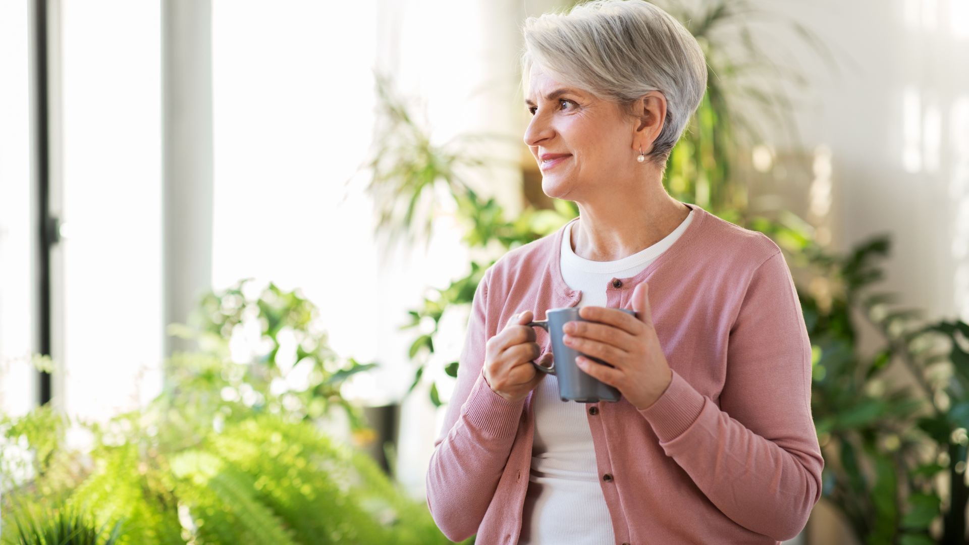 Woman holding a mug by a bright window, wearing a pink cardigan in a plant-filled room