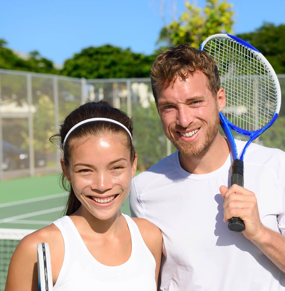 Two tennis players smiling on a tennis court, woman holding a racket, man with a racket over his shoulder.