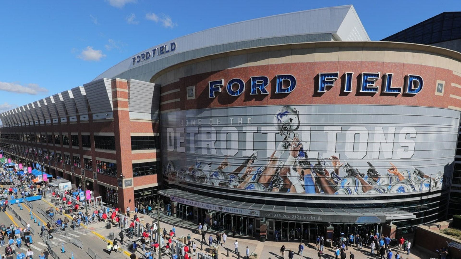 Ford Field stadium exterior with a large Lions banner and crowds outside on a sunny day