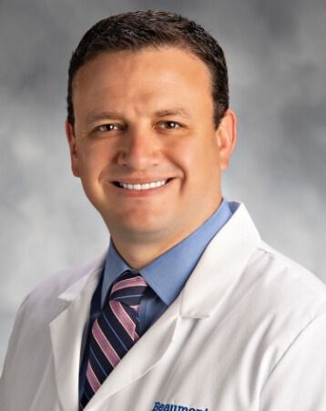 Portrait of a smiling Dr. Joshua Vrabec, MD in a white coat, blue shirt, and striped tie against a gray background
