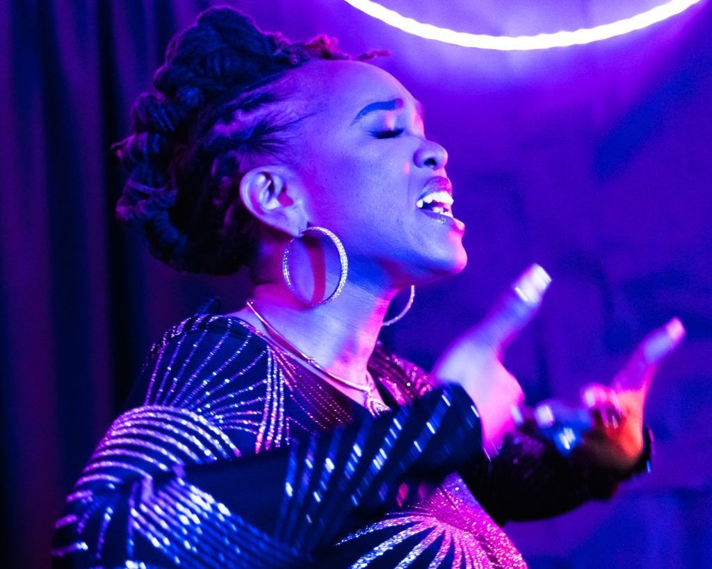 Singer performing on stage under purple neon lights, wearing a striped outfit and large hoop earrings.