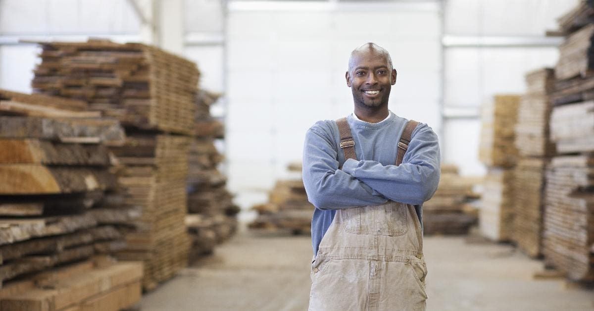 Worker in a warehouse standing with arms crossed, wearing a blue shirt and apron, with stacks of wood behind him.