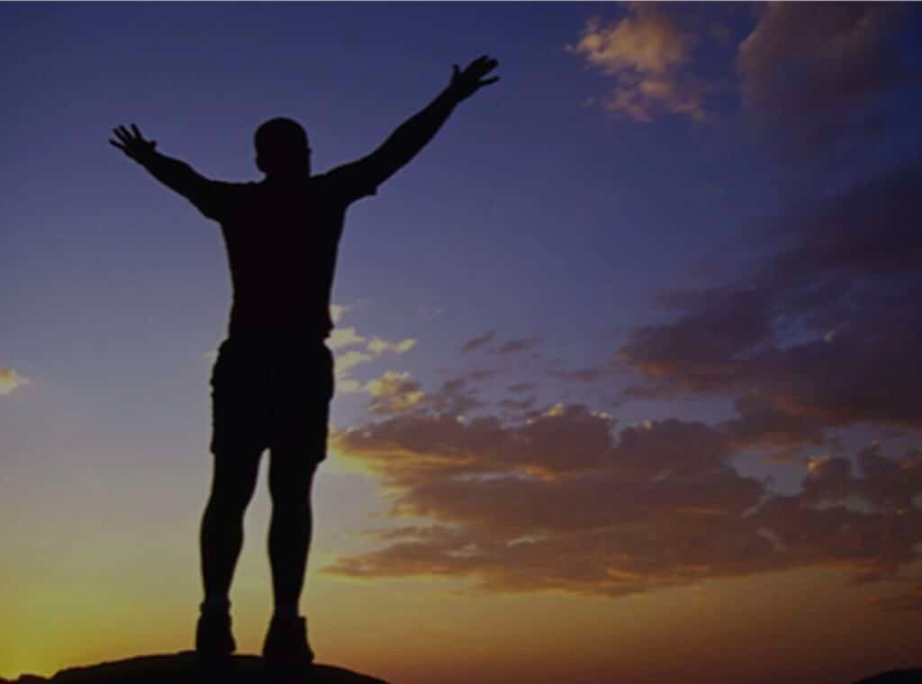Silhouette of a person standing on a hill with arms raised at sunset under a colorful sky