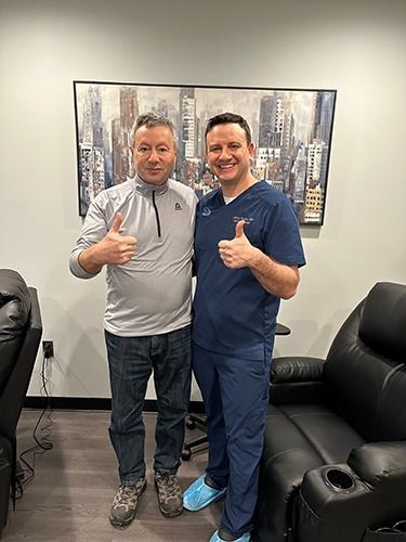 Two men standing in an office, one giving a thumbs-up, with black leather chairs and a cityscape painting behind them.