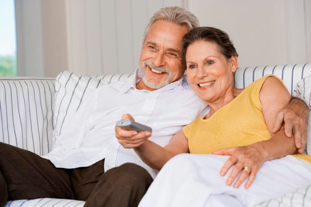 Smiling couple relaxing on a white couch, holding a remote control, in a bright living room