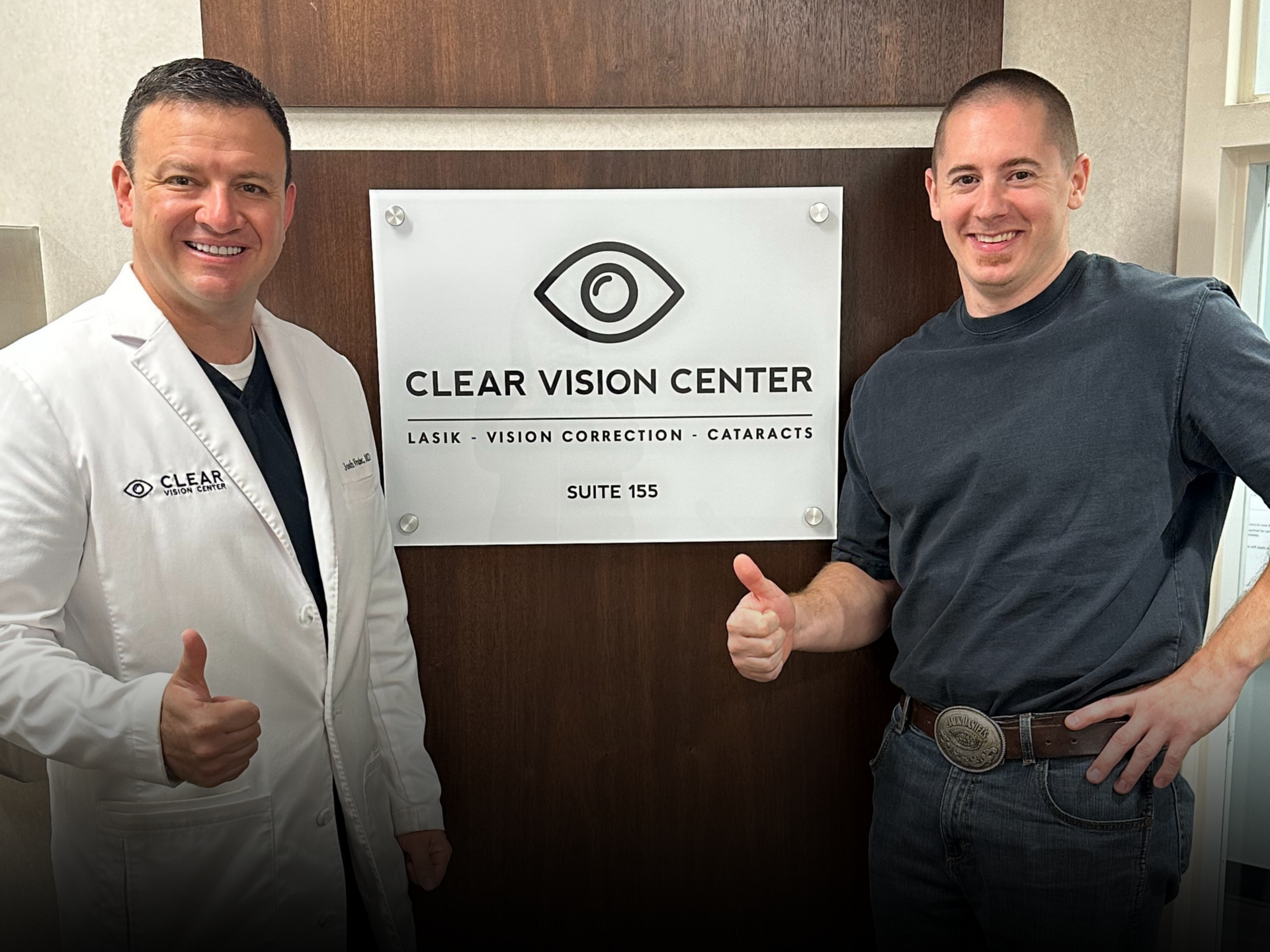 Two men posing with thumbs up beside a Clear Vision Center sign on a wood-paneled wall