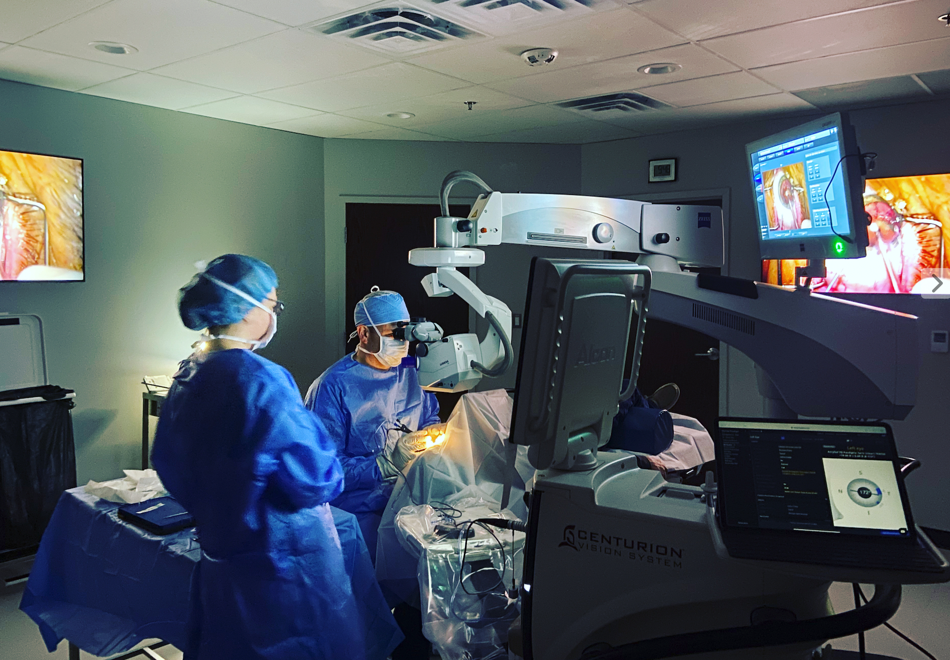 Surgeons in blue scrubs operate in a dim operating room with monitors and bright surgical lights.
