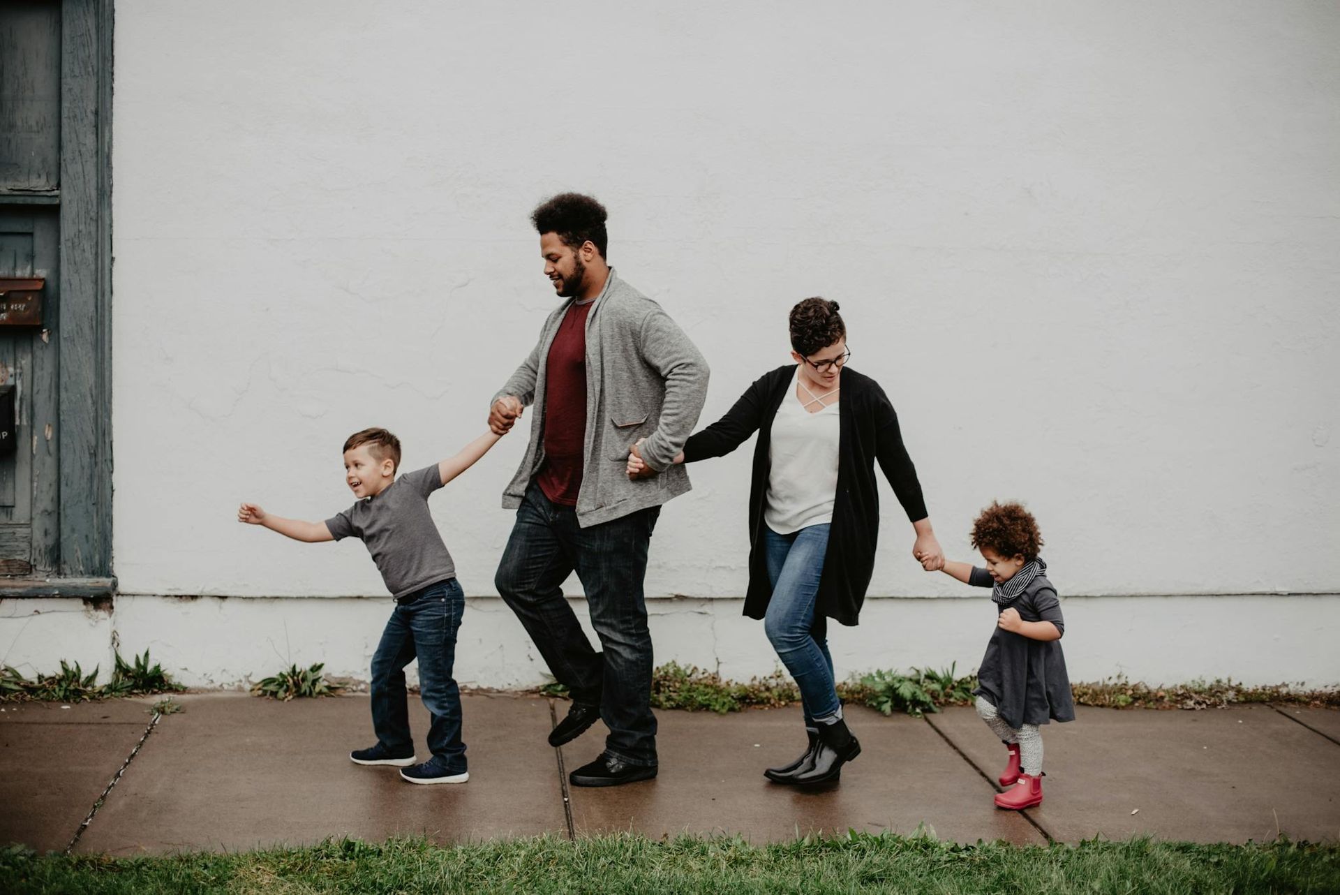 Family of four walking hand in hand along a sidewalk beside a white wall