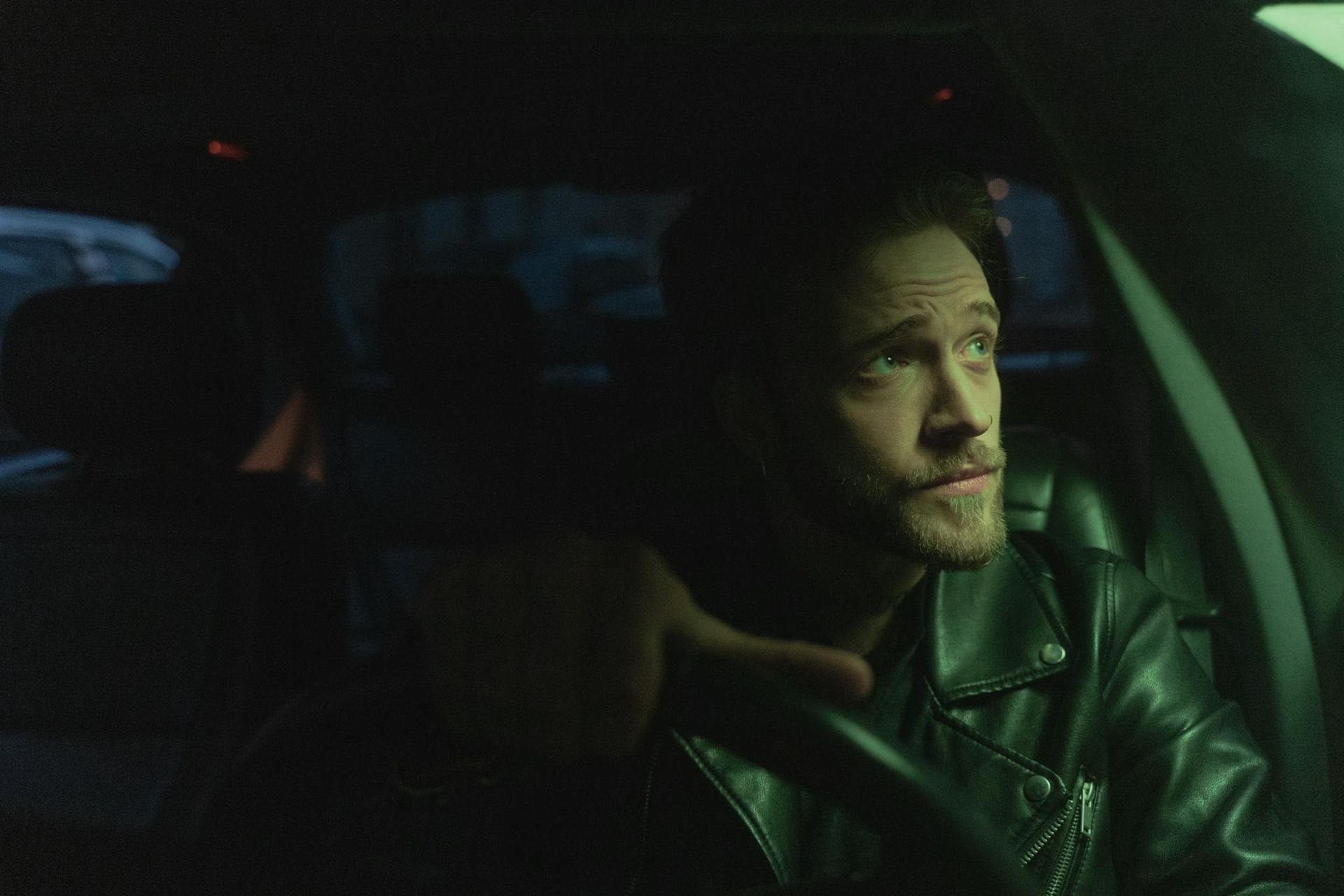 Man in a leather jacket driving at night, lit by green dashboard light