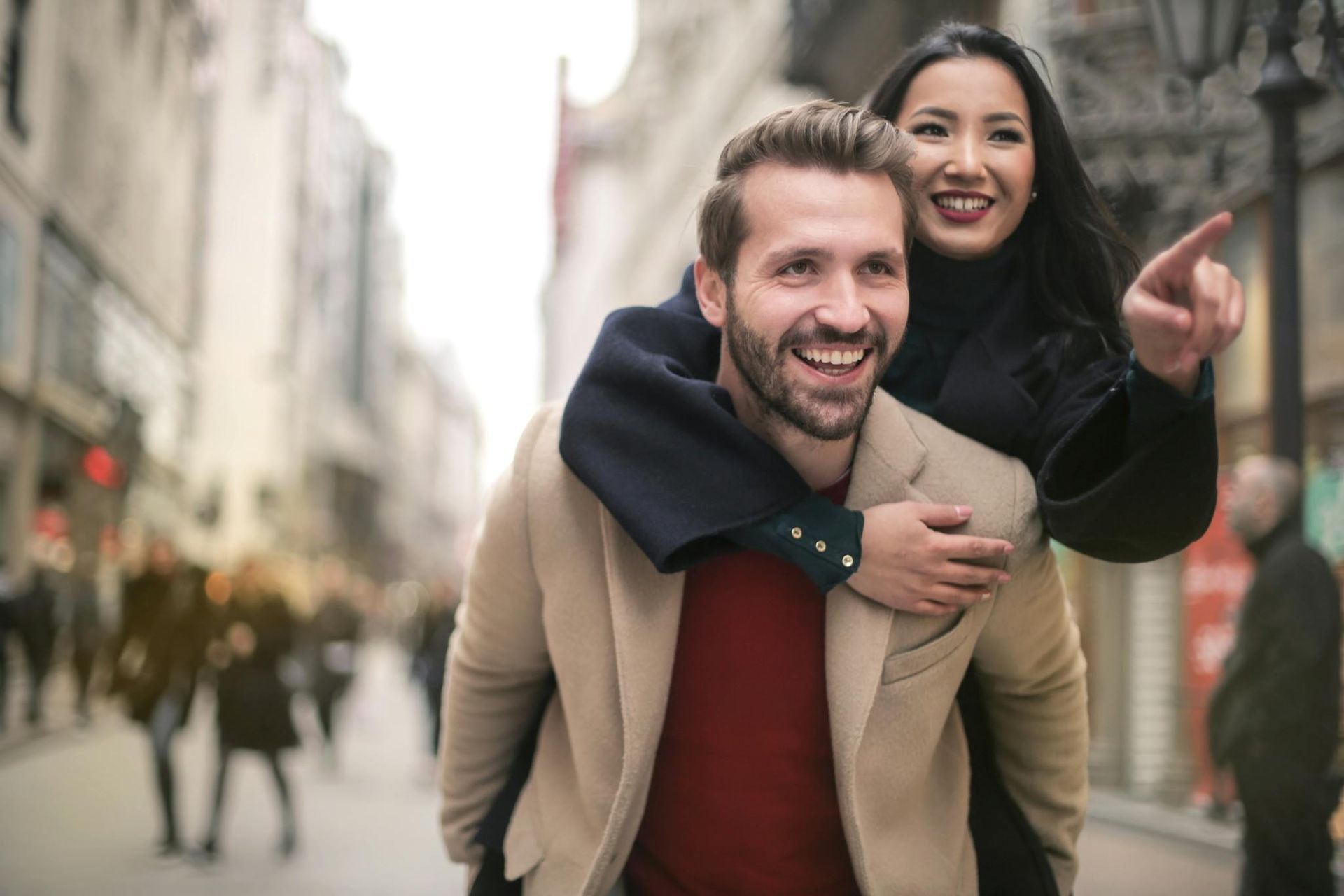 Smiling couple on a city street, woman riding piggyback and pointing ahead, both in winter coats