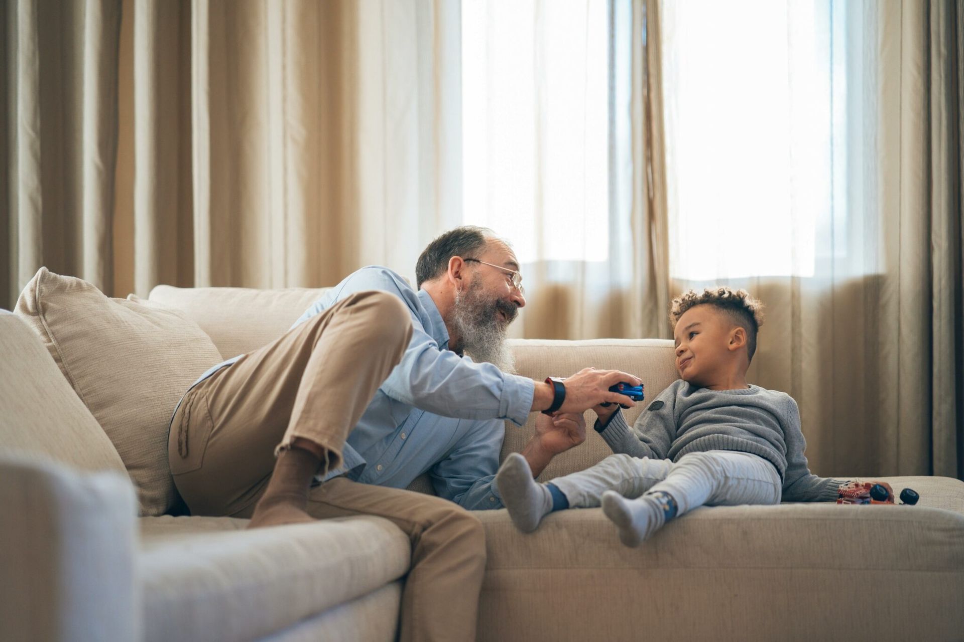 Adult and child on a couch, smiling and playing with a remote in a sunlit living room.