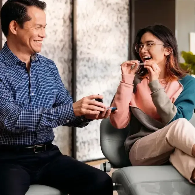Man smiling, handing woman clear aligner trays. She laughs, holding a tray. Interior setting.