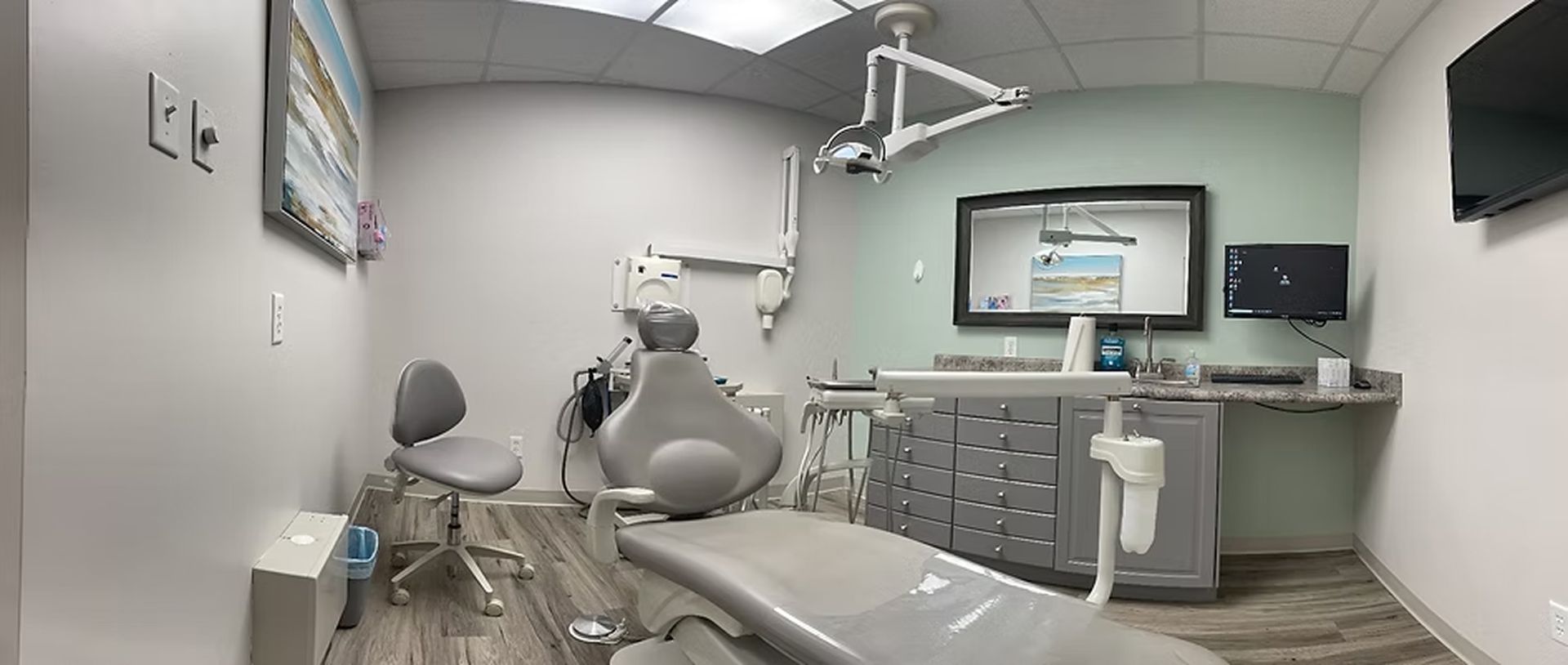 A dentist's office with dental chair, equipment, and cabinets. Light gray and green walls.