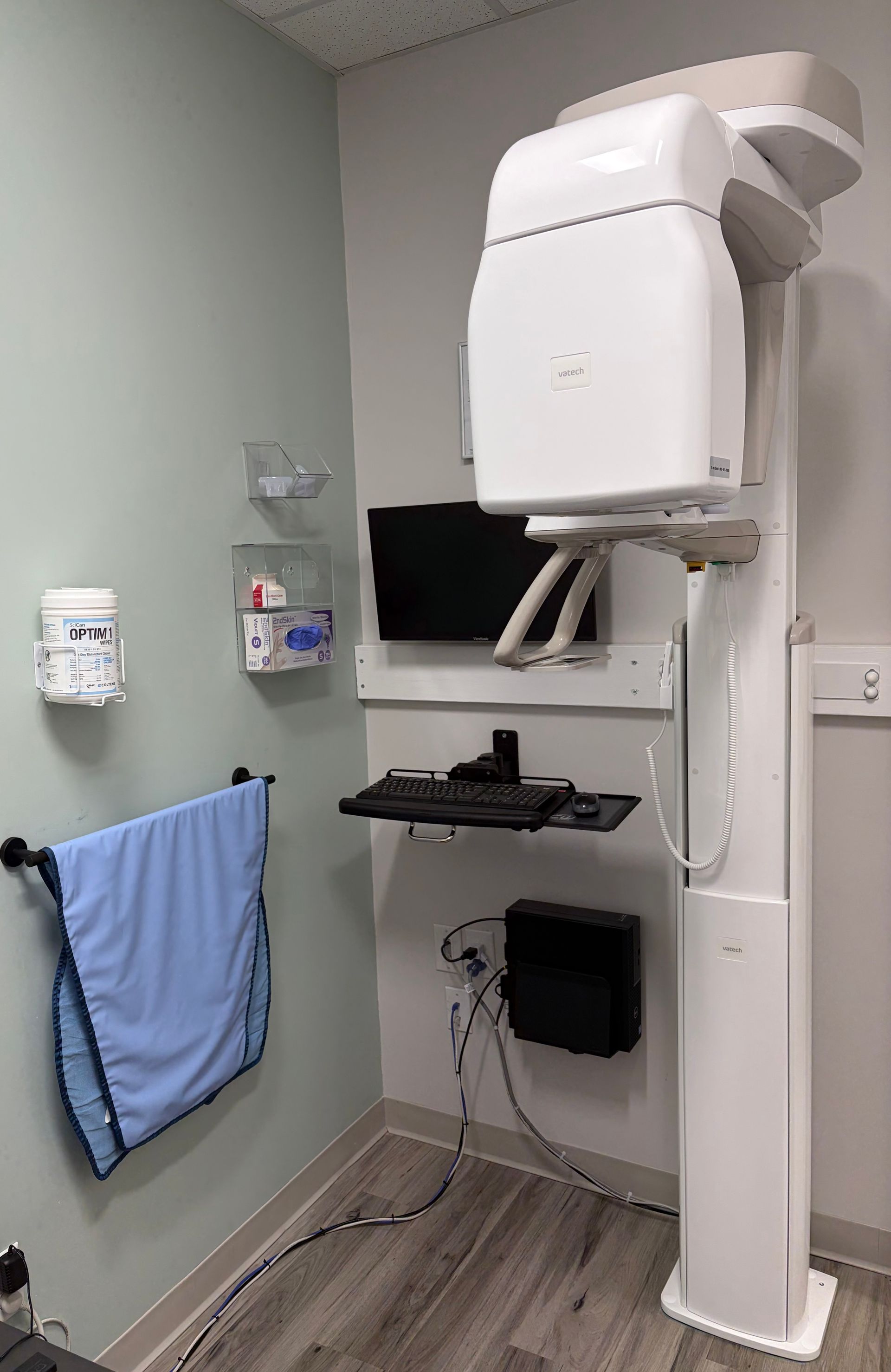 Dental X-ray machine in a corner of a room, next to a wall with a lead apron and supplies.