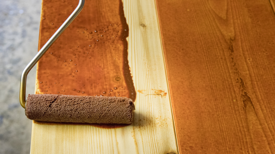 Wooden planks being stained with a roller, showing a color contrast.