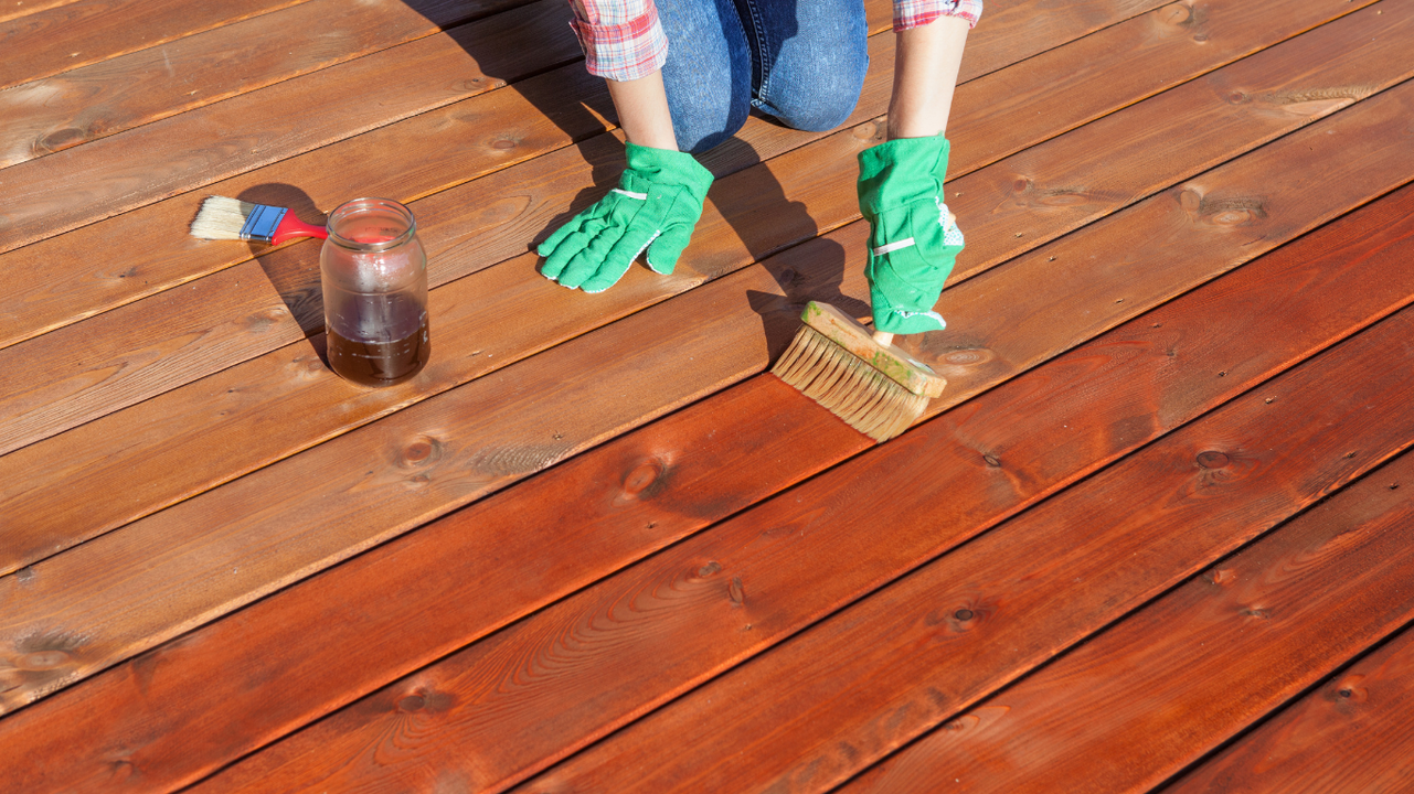 Person staining a wooden deck with a brush, wearing gloves. Half the deck is stained reddish-brown.