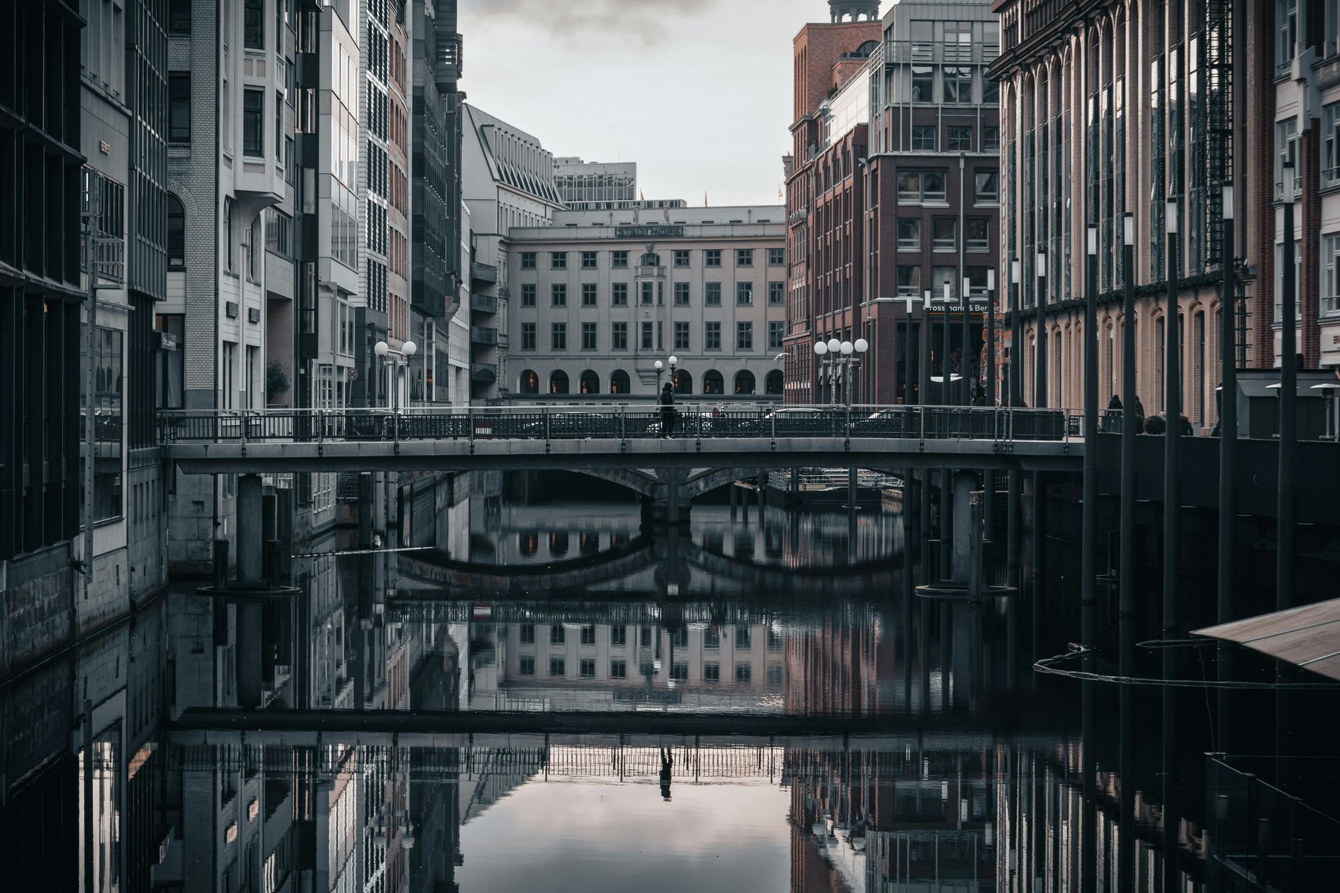 A bridge over water with buildings reflecting in the water. Gray sky, daytime.