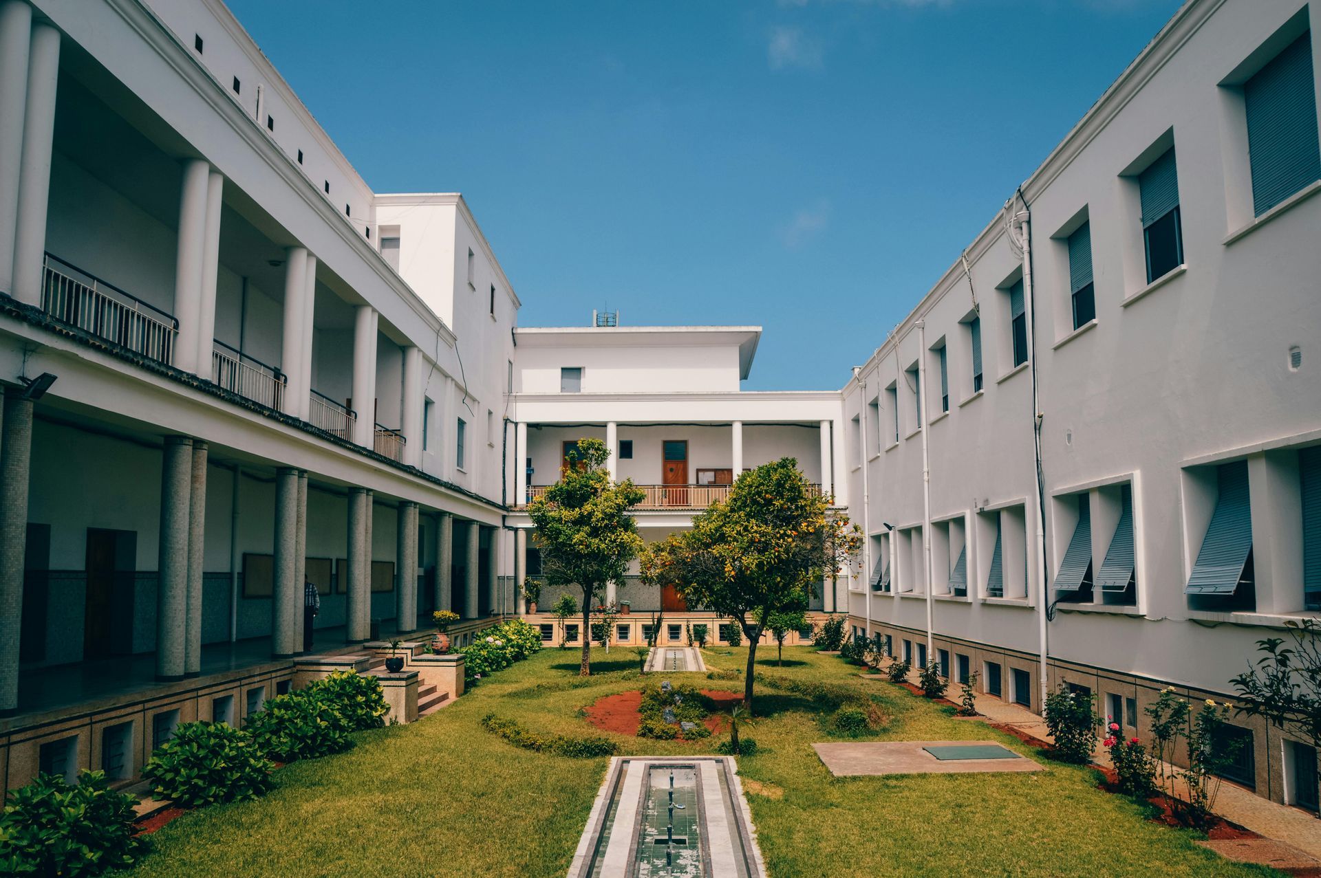 Courtyard with white buildings, green grass, trees, and a fountain under a blue sky.