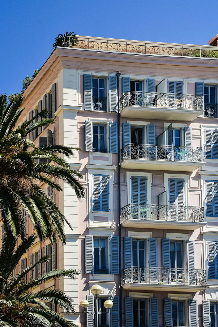 Pink building with blue shutters and balconies under a bright blue sky. Palm tree in foreground.