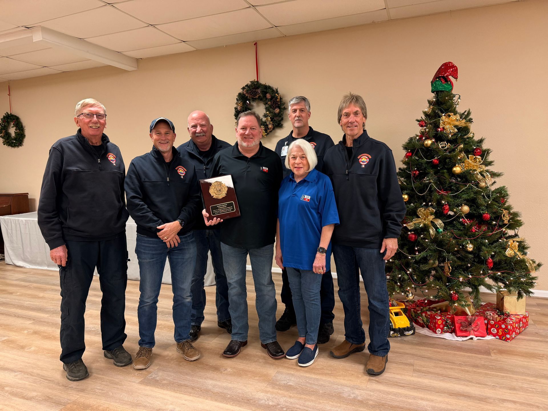 Group of people with an award, standing by a decorated Christmas tree in a room with festive decorations.