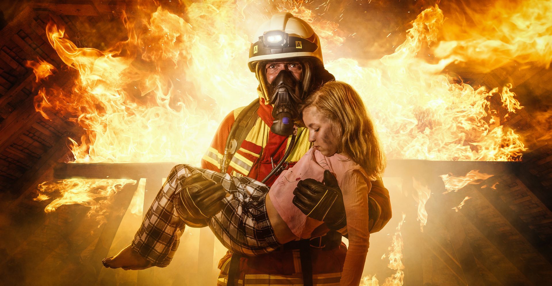 Firefighter carries a child from a burning building. Flames engulf the background.