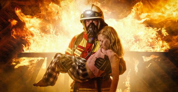 Firefighter carries a child from a burning building. Flames engulf the background.