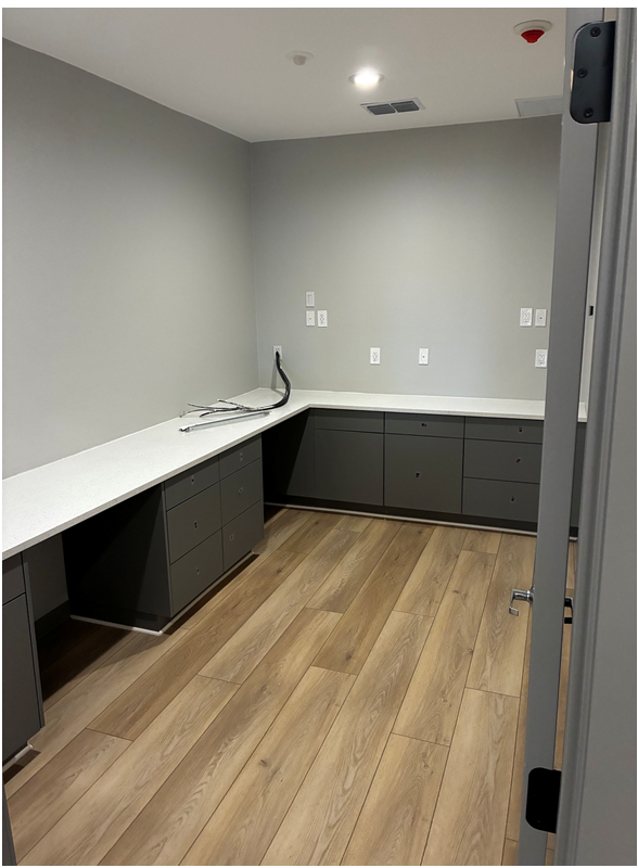 An L-shaped workspace with gray cabinets and white countertops against light gray walls and wood-look flooring.
