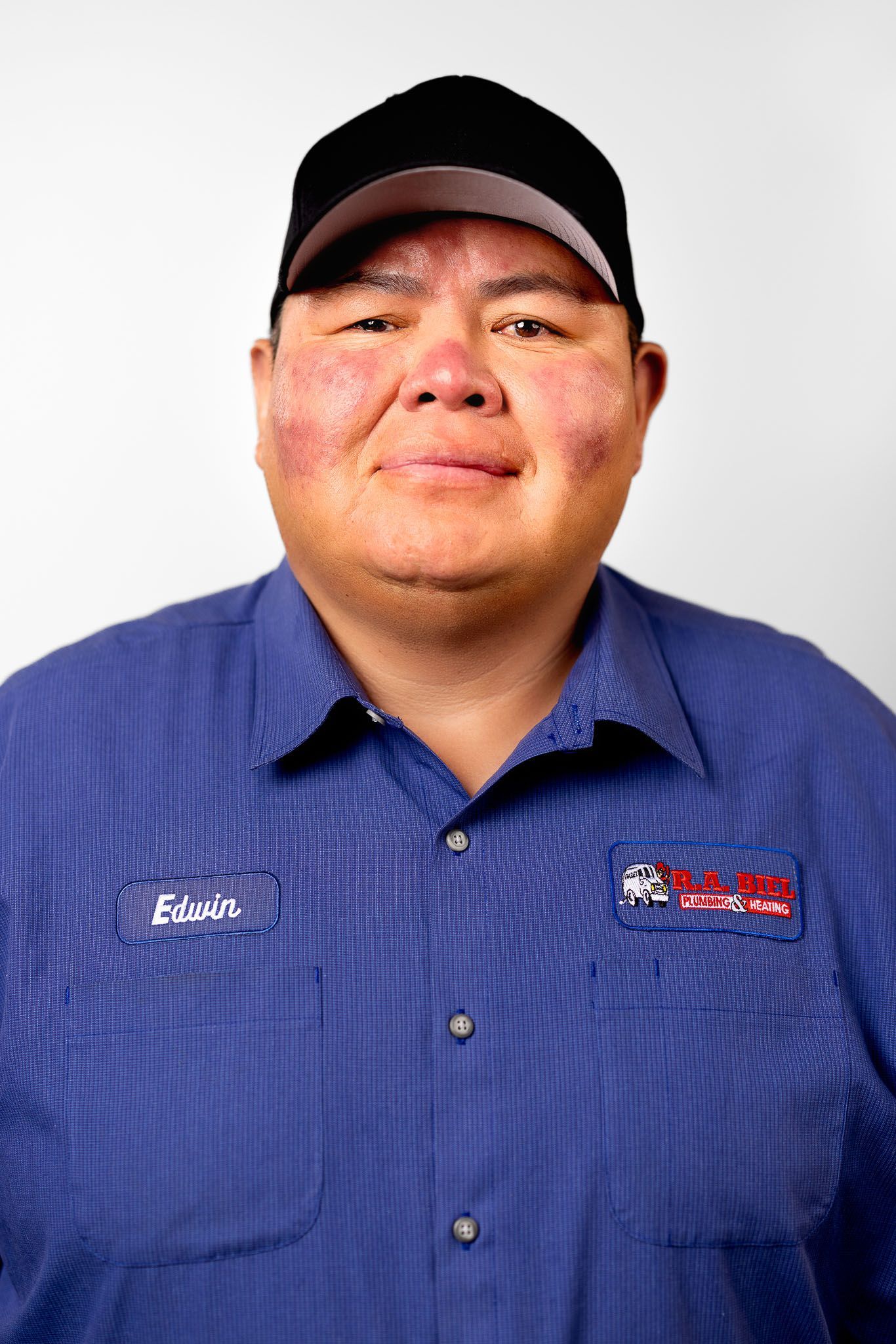 A man wearing a blue shirt and a black hat is standing in front of a white background.