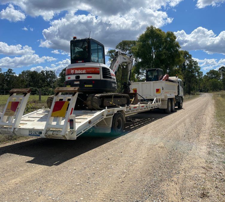 A Bobcat Excavator is Being Transported on a Trailer on a Dirt Road — Upgrade Earthworks In South Kolan, QLD