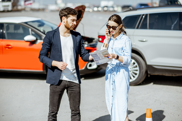 Man gestures near a car with its hood open, while woman on phone reads papers at a parking lot after an accident.