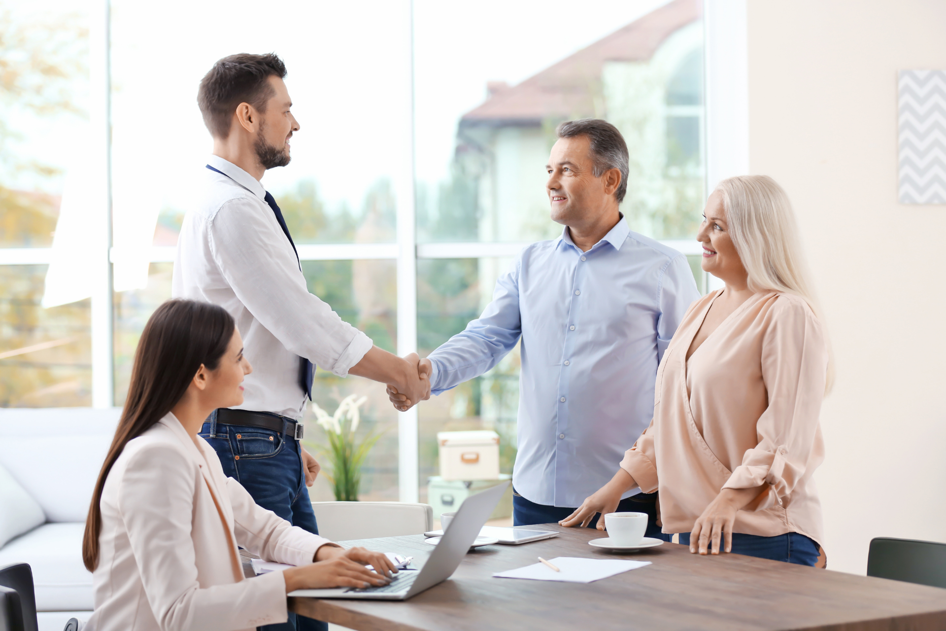 A real estate agent shakes hands with a couple; another agent types on a laptop.