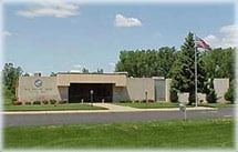 Tan brick building with a dark entryway, American flag, and surrounding trees.