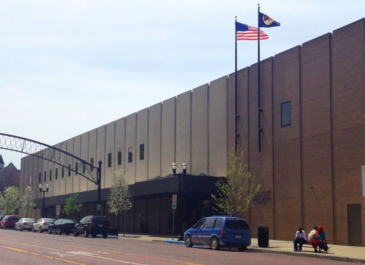Brick building with US and state flags flying; cars parked on street; people on bench.