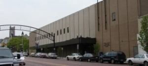 A street with cars parked in front of a long, tan building with vertical lines.