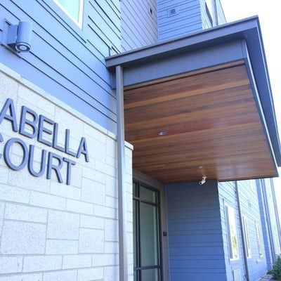 Entrance to Isabella Court apartment building, featuring a wooden canopy, light blue siding, and a sign.