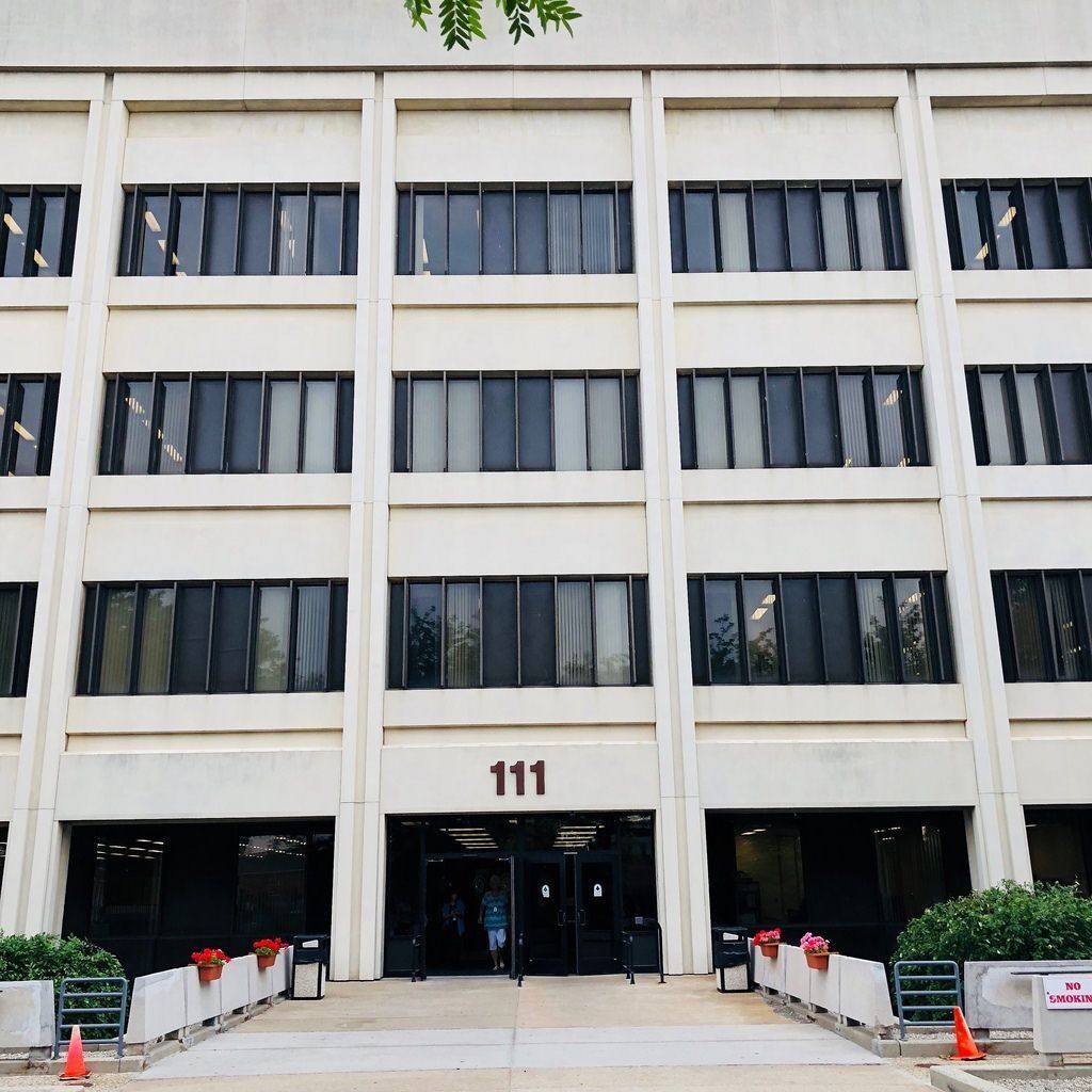 Three-story concrete building with many dark rectangular windows; entrance with 111 on top; bushes and small barriers.