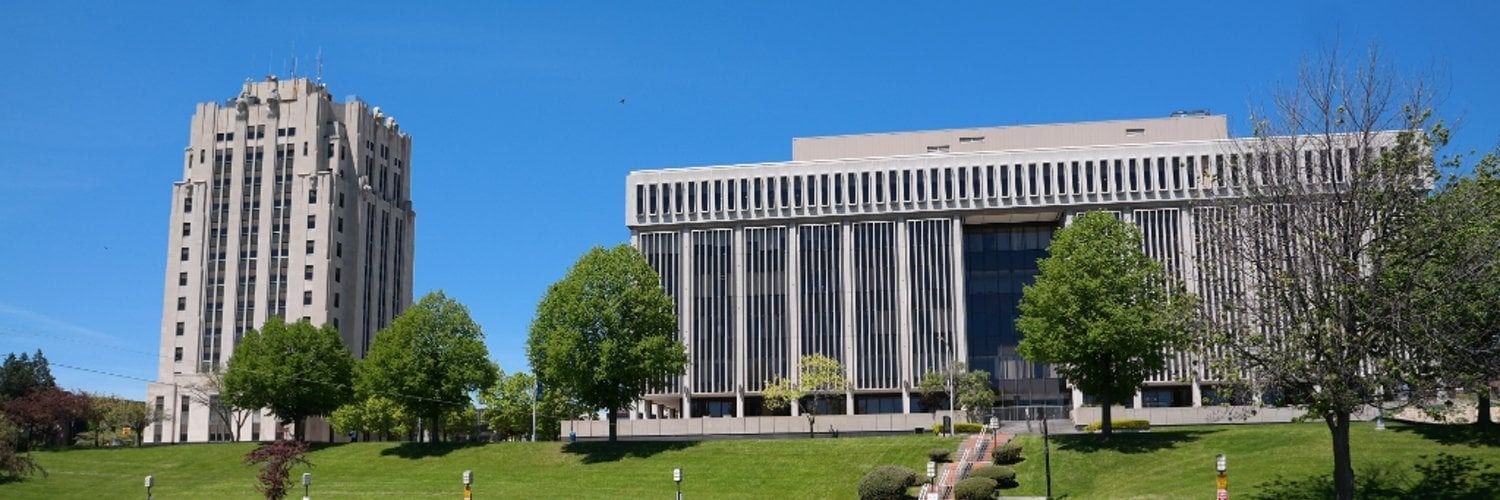 Buildings on a grassy hill under a blue sky, with trees in front of the buildings.