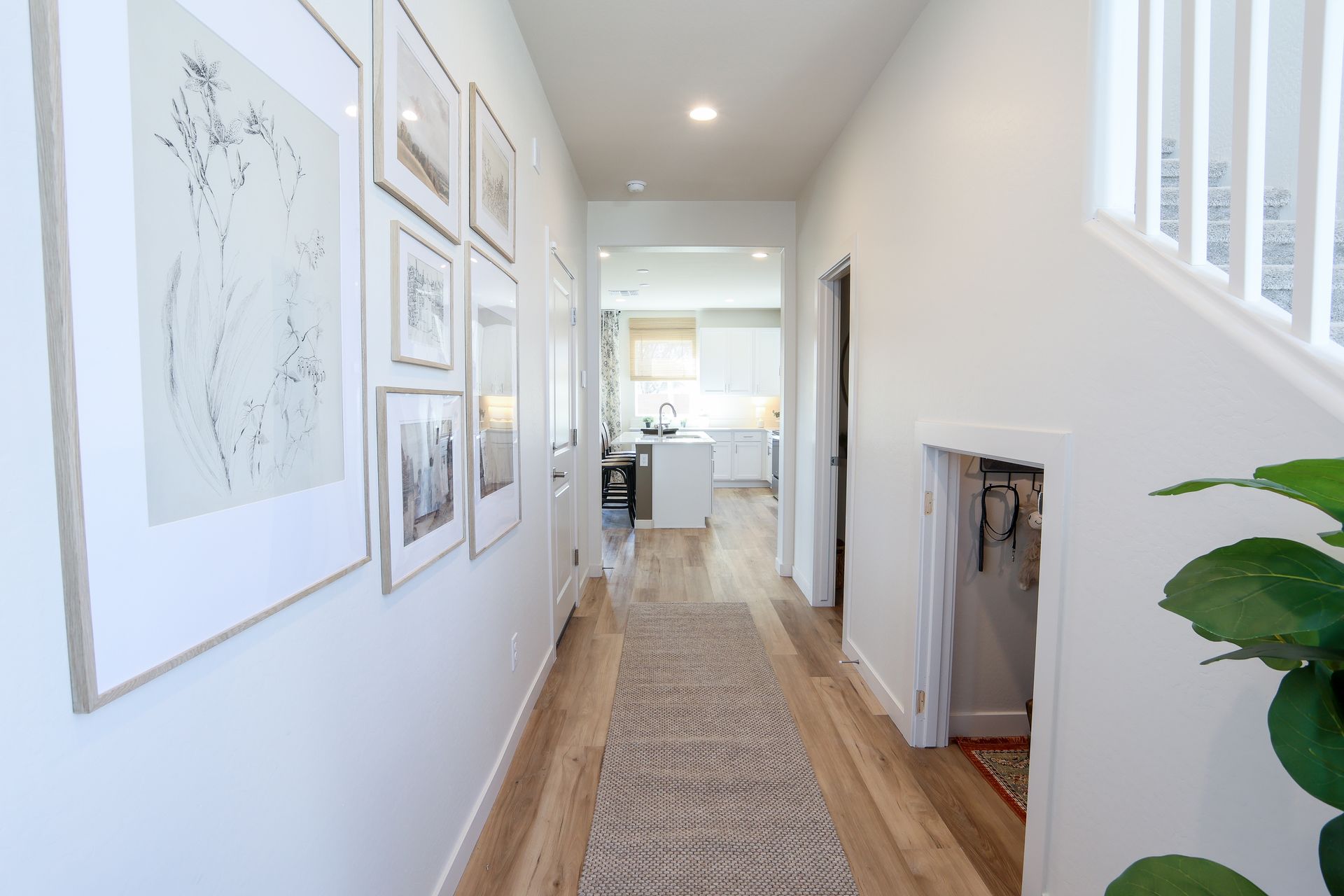 Bright hallway with framed art, white walls, wood floors, runner rug, and a glimpse of a kitchen at the end.
