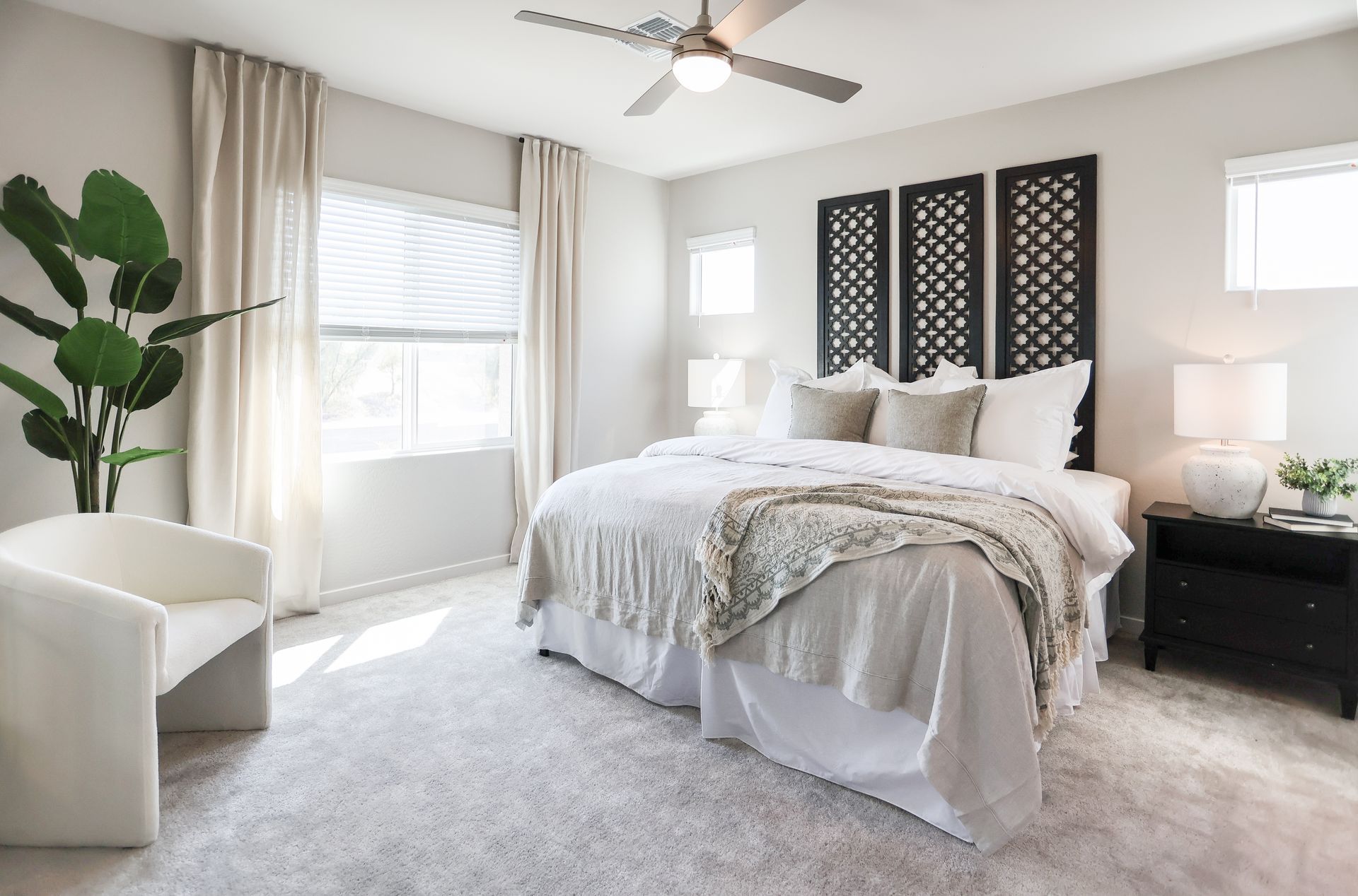 Bright bedroom with a bed, white chair, nightstands, and large windows with curtains.